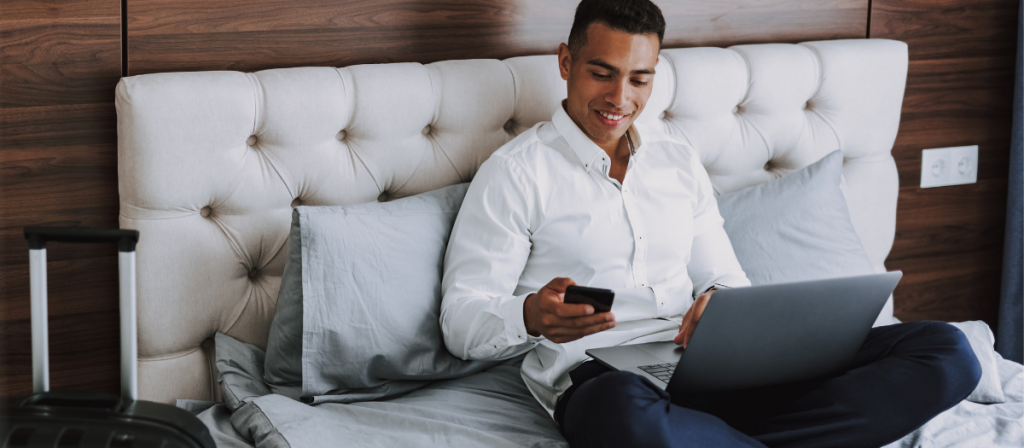 Male business traveler sitting on bed looking at his cellphone with a laptop in his lap.