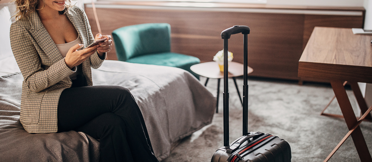 Female business traveler sitting on a hotel bed, smiling at her cell phone.