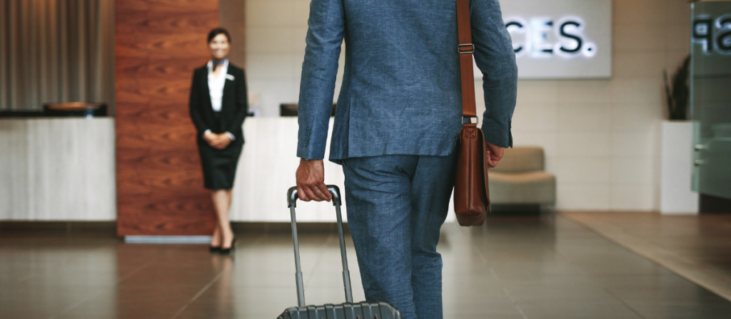 A man in a business suit walks toward the reception desk of a hotel to check in. A receptionist waits in the background.